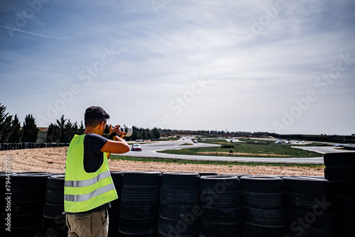 Young, tanned guy in reflective vest captures racing cars with wide-angle lens at Burgos track.