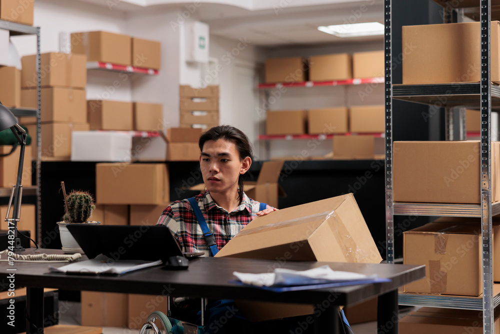 Warehouse employee in wheelchair processing order on laptop while holding cardboard box ready for shipment. Storehouse asian young man delivery manager working in disability friendly workplace Warehouse employee in wheelchair processing order on laptop while holding cardboard box ready for shipment. Storehouse asian young man delivery manager working in disability friendly workplace