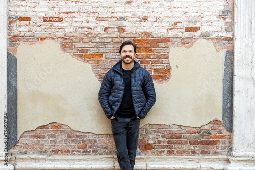 An elegant bearded man wearing casual clothes stands smiling leaning against wall with hands in his pockets in Venice, Italy. Old venetian grungy brick wall with damaged plaster