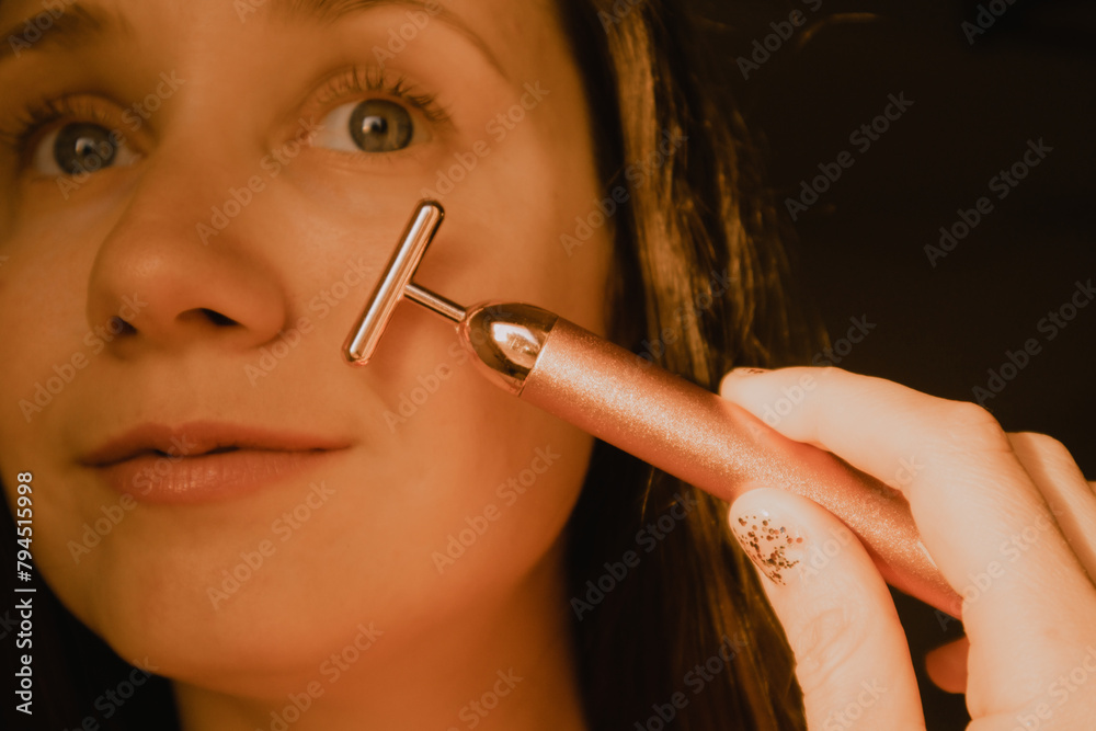 Obraz premium Young beautiful white woman, girl is using vibrating golden facial massager on black background. Face massage. Natural beauty youth Female skin care.