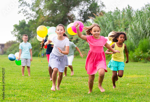 Multiracial group of cheerful tweenagers holding colored balloons in hands, running together in summer city park. Happy childhood concept