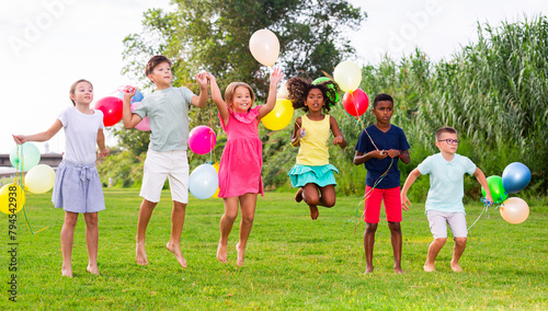 Portrait of cheerful tween girls and boys holding colorful balloons in hands jumping together in summer city park. Concept of happy preadolescence photo