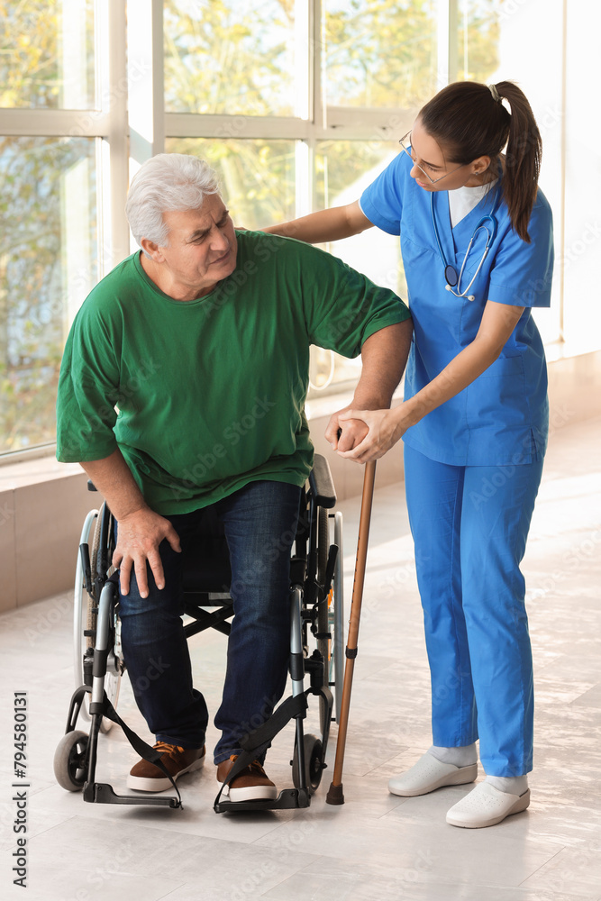 Nurse helping senior man with stick to stand up from wheelchair at hospital