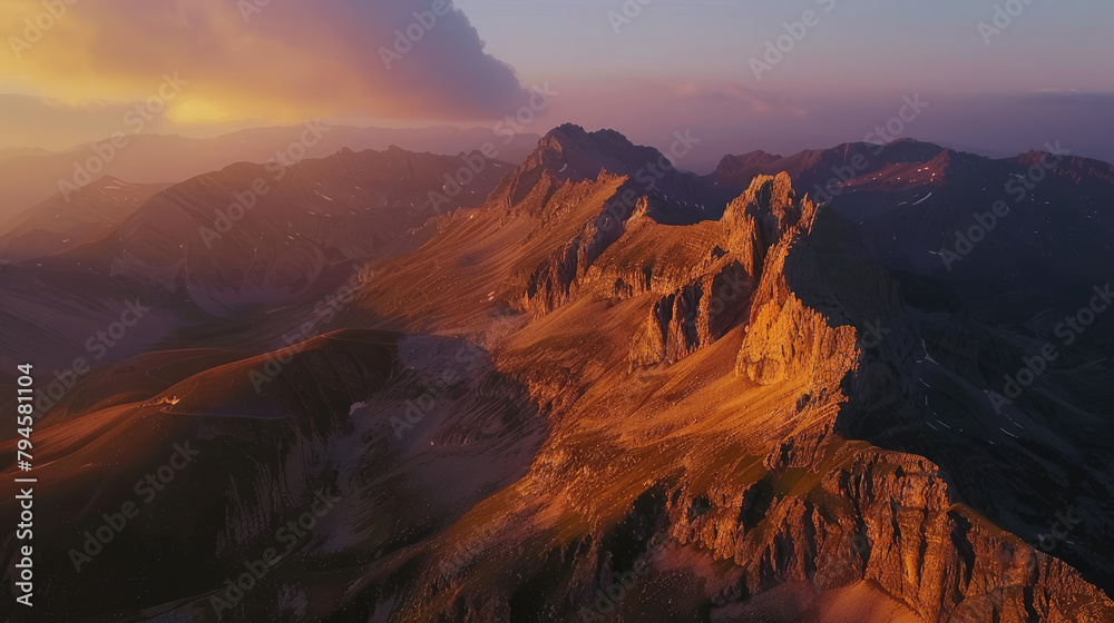 Obraz premium Aerial view of a mountain range at sunrise, the peaks illuminated with golden light against the shadowed valleys