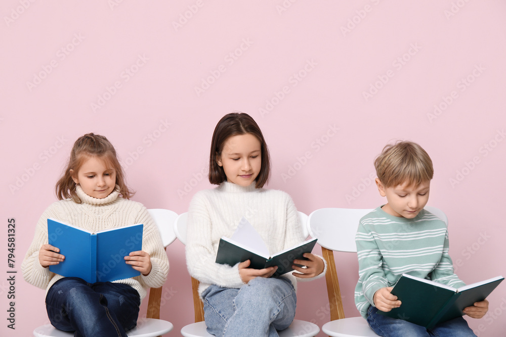 Little children reading books while sitting on chairs against pink background Little children reading books while sitting on chairs against pink background