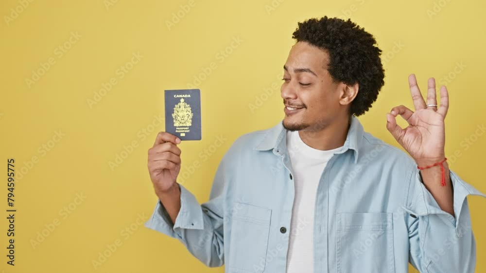 Grinning african american young man joyfully flashes a canadian passport, doing the 'okay' sign with fingers against an isolated yellow wall, radiating confidence and friendly positivity. Grinning african american young man joyfully flashes a canadian passport, doing the 'okay' sign with fingers against an isolated yellow wall, radiating confidence and friendly positivity.