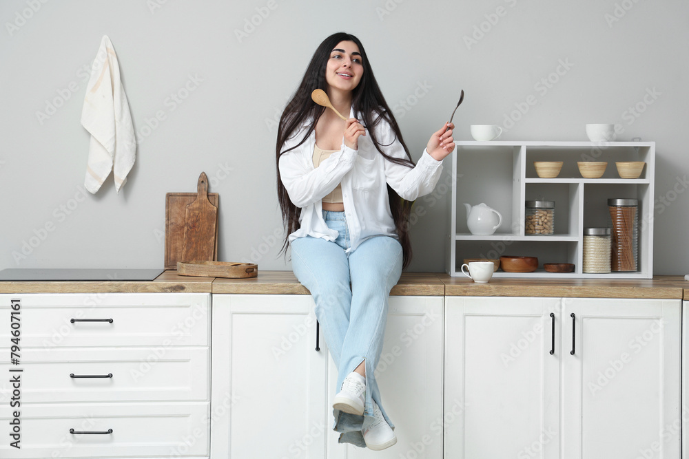 Beautiful young happy woman with wooden spoons sitting on table in kitchen at home