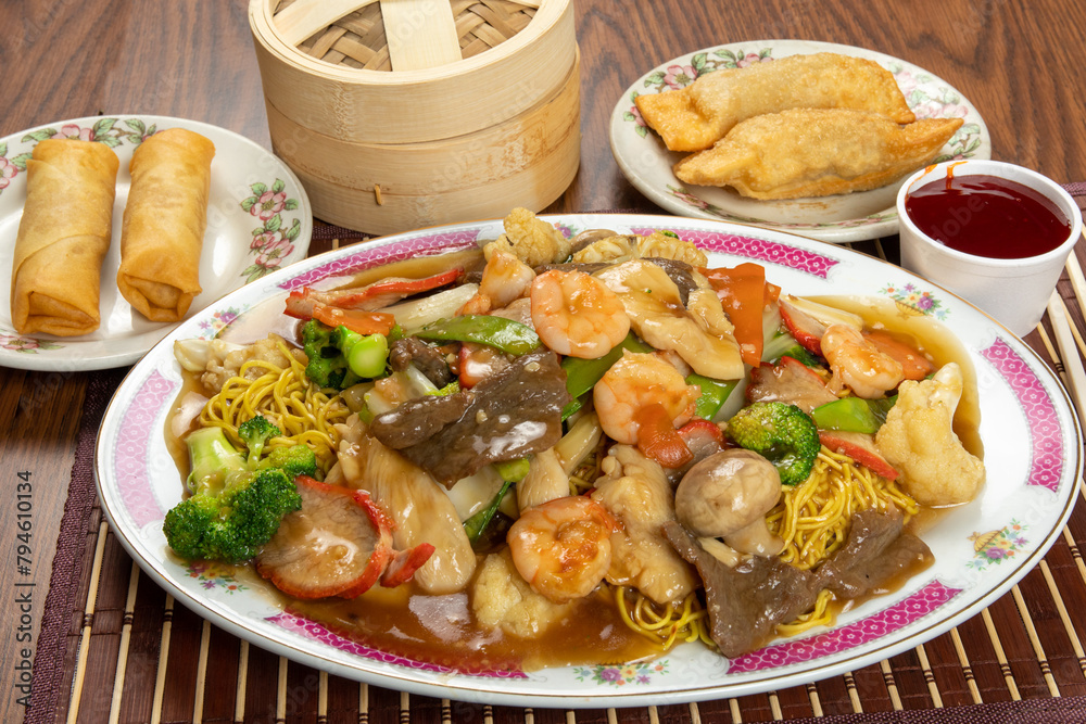 A plate of Cantonese chow mein chicken with plates of egg rolls and spring rolls in the background