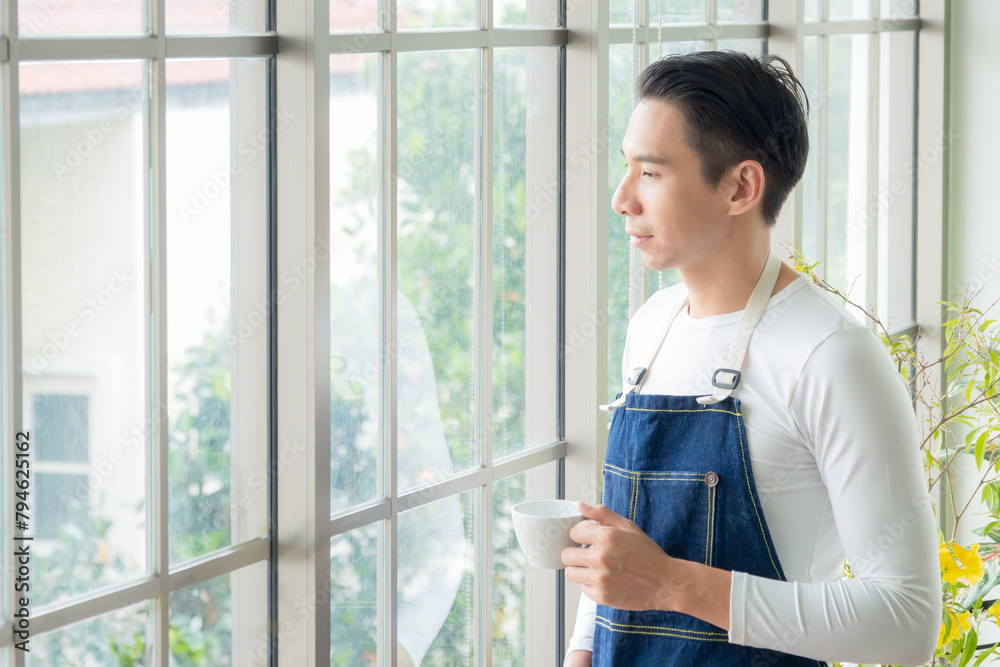 Portrait gardener adult young Asian man one person handsome standing think look and Smile relax happy hand holding drinking cup coffee in room cafe with glass window and green tree in morning time. Portrait gardener adult young Asian man one person handsome standing think look and Smile relax happy hand holding drinking cup coffee in room cafe with glass window and green tree in morning time.