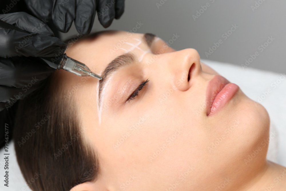 Beautician making permanent eyebrow makeup to young woman on grey background, closeup