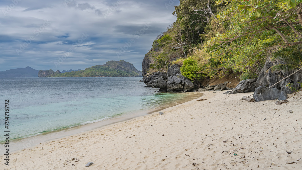 A secluded sandy beach on a tropical island. A calm turquoise ocean. Green vegetation on the coastal cliffs. Mountains in the distance against a blue sky and clouds. Philippines.  Palawan. El Nido.