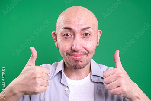 Bald asian man showing two thumbs up gesture and smiling to camera over green background