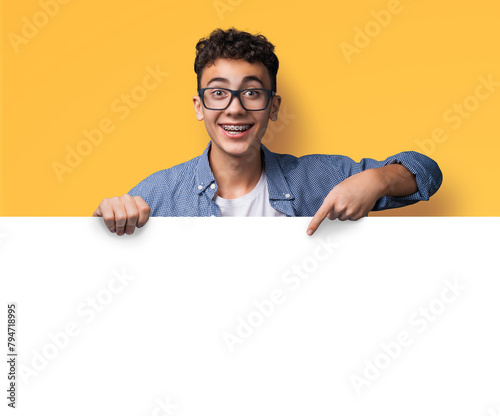 Excited happy young man wear glasses, braces stand behind, peep empty banner mockup sign board signboard, advertise show point sale slogan text place, isolated yellow background. Dental care ad