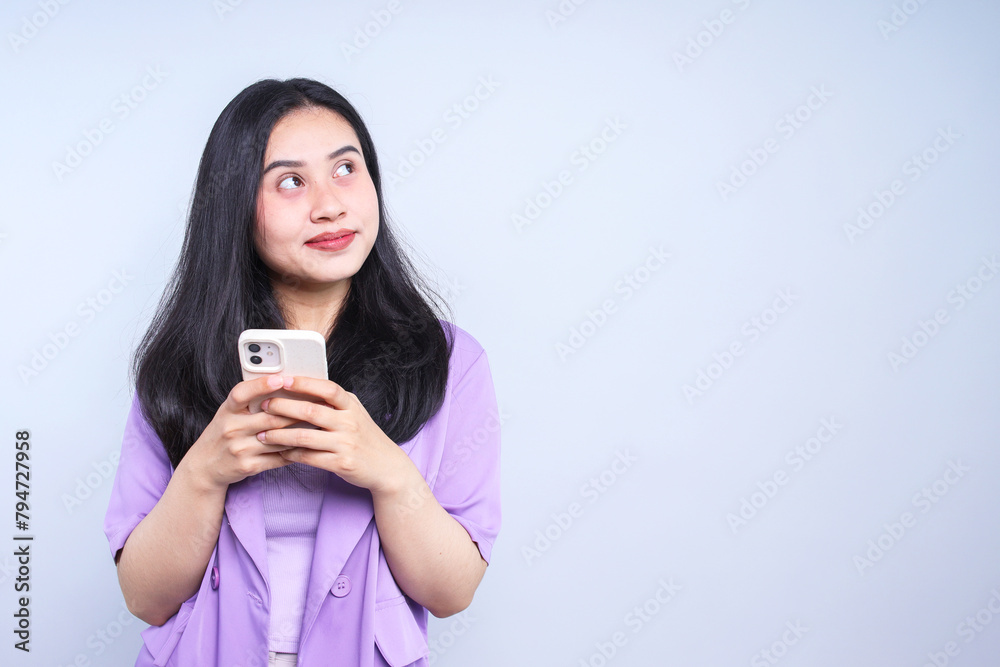Happy Asian woman holding smartphone and looking up over grey background Happy Asian woman holding smartphone and looking up over grey background