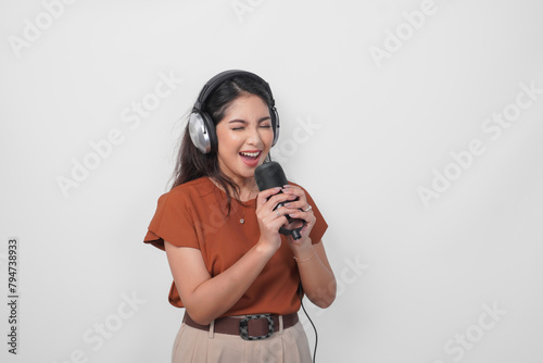 Overjoyed young woman wearing brown shirt and headphone to listen music and singing along isolated over white background. (ID: 794738933)