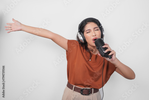 Overjoyed young woman wearing brown shirt and headphone to listen music and singing along isolated over white background. (ID: 794739366)