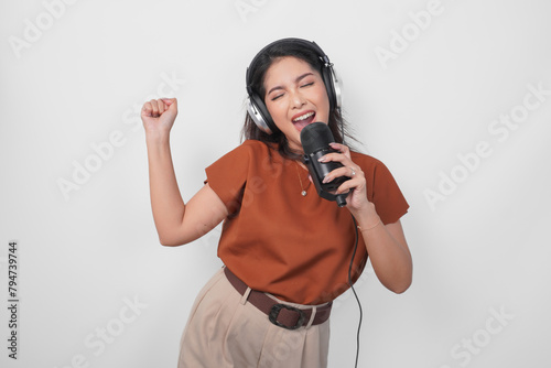 Excited young woman wearing brown shirt and headphone to listen music and singing along to the microphone isolated over white background. (ID: 794739744)