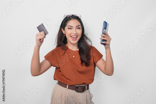 Happy successful Asian woman wearing brown shirt is holding her smartphone and credit card over isolated white background.