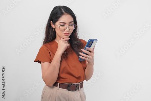 Thoughtful young Asian woman wearing brown shirt and eyeglasses looking aside while holding smartphone isolated over white background.