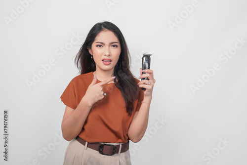 Young Asian woman wearing brown shirt pointing to an electric shaver she holds with confused expression, isolated by white background.