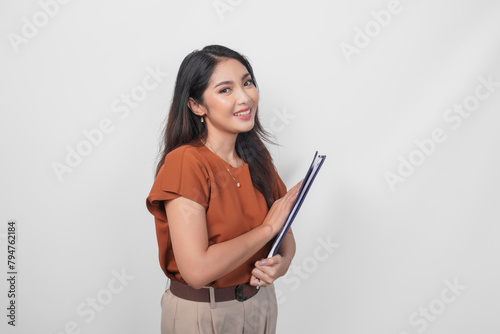 Beautiful young Asian woman wearing brown shirt smiling while holding document over isolated white background.