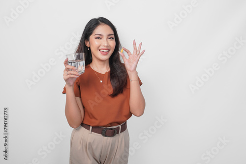 Smiling Asian woman in brown shirt drinking medicine pill with a glass of water to stay healthy isolated by white background.
