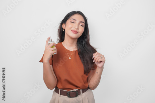 Beautiful young Asian woman in brown shirt holding bottle of perfume and spraying on her neck isolated by white background. (ID: 794764985)