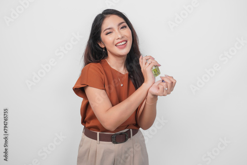 Smiling young Asian woman in brown shirt holding bottle of perfume and spraying on her wrist isolated by white background. (ID: 794765755)
