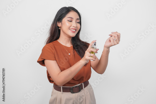 Smiling young Asian woman in brown shirt holding bottle of perfume and spraying on her wrist isolated by white background. (ID: 794765789)