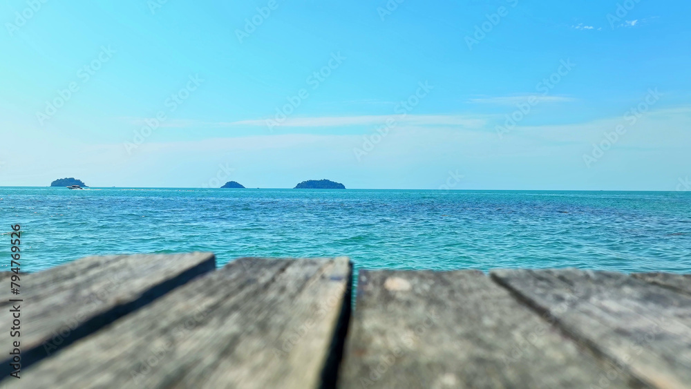 A tropical paradise: Crystal-clear turquoise waters stretch out beneath a radiant blue sky, with an enchanting island on the horizon and a wooden bridge in the foreground. Ko Chang island, Thailand. 
