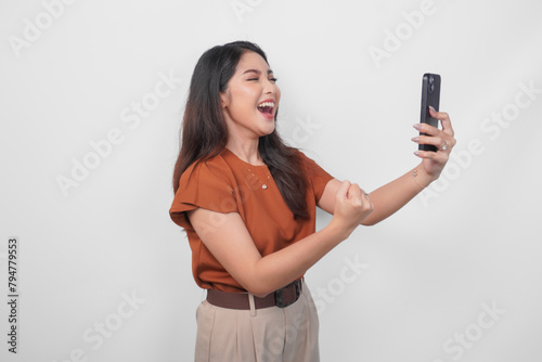 Joyful young Asian woman in brown shirt doing video call with happy successful expression and clenched fist gesture isolated over white background.