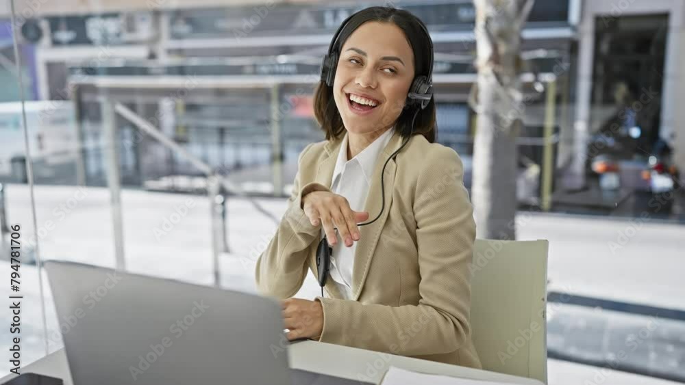 A smiling, young hispanic woman wearing a headset and beige blazer sits at a desk against a blurred office backdrop. A smiling, young hispanic woman wearing a headset and beige blazer sits at a desk against a blurred office backdrop.
