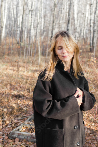 A young woman wearing a black coat stands pensively in a sparse autumn forest. The trees are bare, and the leaves are scattered on the ground. She looks off into the distance, lost in thought.