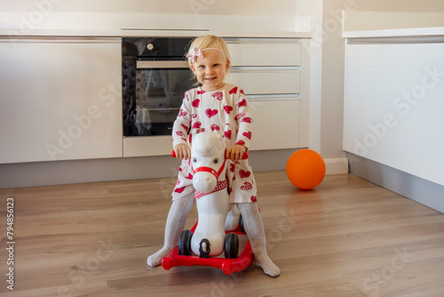 Children play indoors. Children ride a toy rocking horse. A little blonde girl in a dress is playing in the kitchen. A beautiful nursery for kids. A cute kid is having fun at home.