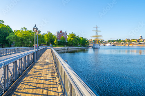 A vibrant day at Skeppsholmen Bridge in Stockholm, with clear skies and sunlight bathing the walkway, the calm water, and historic buildings in the distance. Stockholm, Sweden photo
