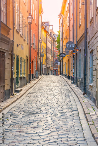 A narrow, sunlit cobblestone street lined with colorful historic buildings in the old town of Gamla Stan, Stockholm, Sweden, on a clear day.