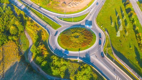 Aerial perspective of a busy roundabout with multiple exits, surrounded by lush green trees and landscape, bathed in sunlight.