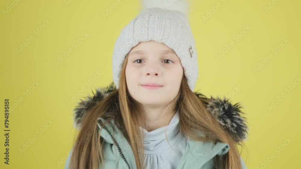 A beautiful little girl dressed in a green down jacket and adjusting her hat looks and smiles at the camera on a yellow background. A beautiful little girl dressed in a green down jacket and adjusting her hat looks and smiles at the camera on a yellow background.
