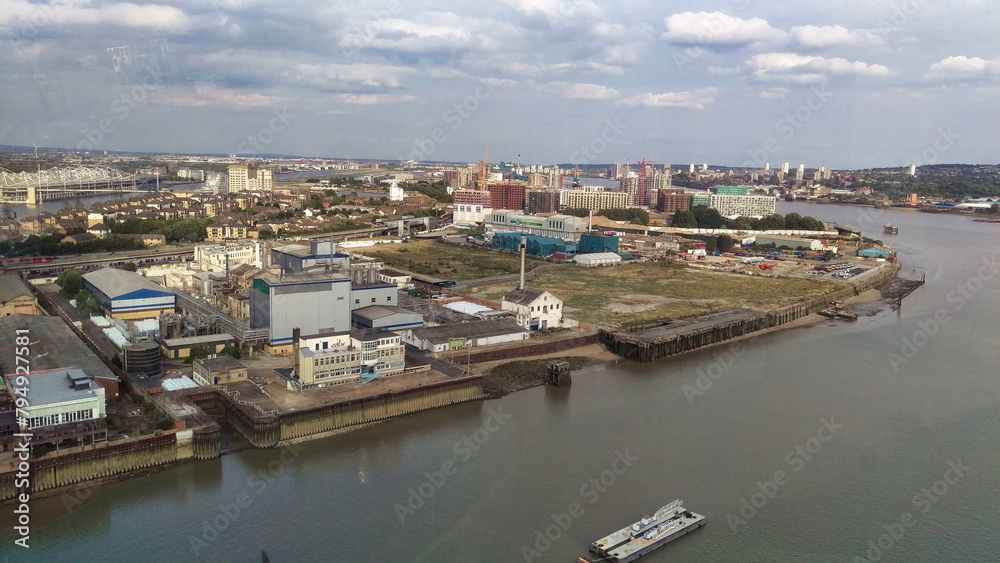 Panoramic view of an industrial riverside with warehouses, multiple buildings, and cranes against a cloudy sky.