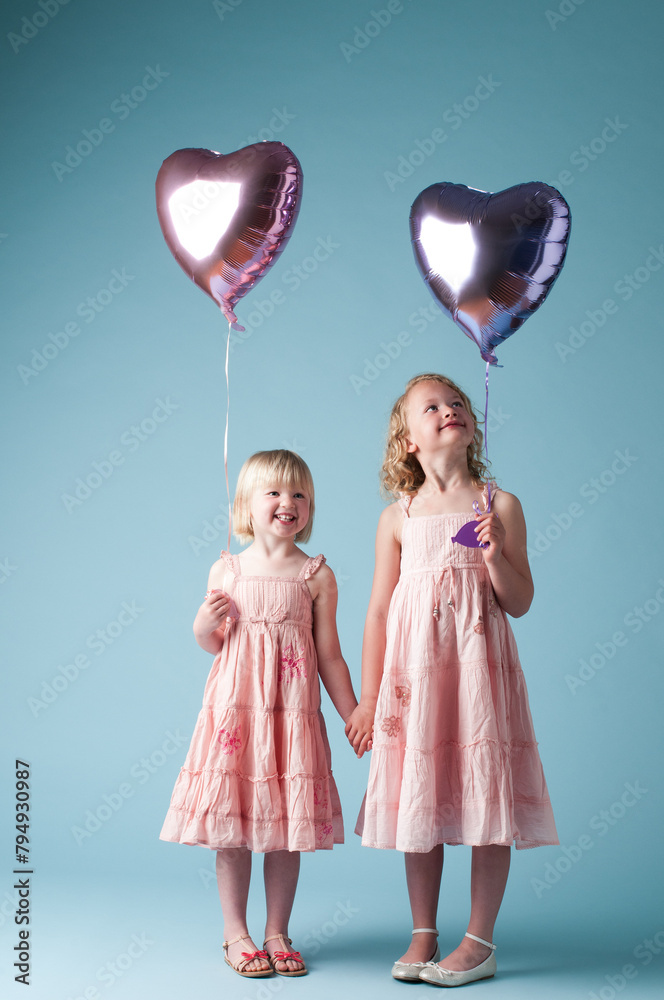 Children, holding hands and sister with heart balloon in studio on blue background for family or party. Birthday, celebration event or happy with smile of girl kids on color backdrop together Children, holding hands and sister with heart balloon in studio on blue background for family or party. Birthday, celebration event or happy with smile of girl kids on color backdrop together