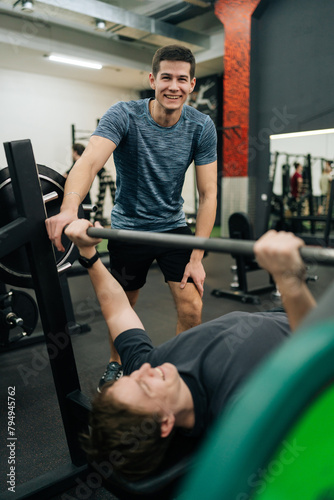 Cheerful experienced fitness instructor helping smiling beginner sportsman doing barbell bench press exercise during personal workout in gym. Male doing barbell bench press under couch supervision