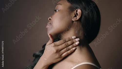 Side view of beautiful Black woman with shiny hair standing in studio and applying moisturizing cream to neck while taking care of skin