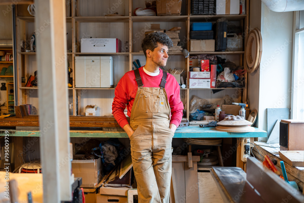 Relaxed thoughtful man carpenter take break after DIY work in joiner workshop. Dreaming craftsman resting looking to window thinking about new handmade project standing with hands in pockets coveralls