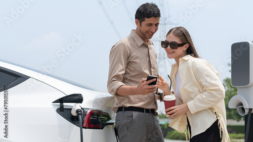 Young couple with coffee pay for electricity while recharge EV car battery at charging station connected to electrical power grid tower as electric industrial for eco friendly car travel. Expedient photo