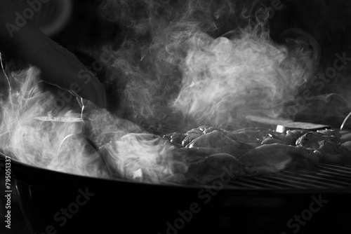 visuale in primo piano, in bianco e nero e su sfondo scuro che mostra vari pezzi di carne mentre vengono cucinati sulla griglia ed emettono fumo photo
