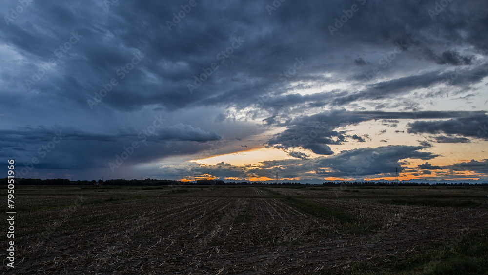 inquadratura panoramica con vista sul paesaggio naturale di campagna nel nord est Italia, di sera, al tramonto, sotto un cielo nuvoloso e temporalesco, con sfumature di colore tra l'arancione e il blu