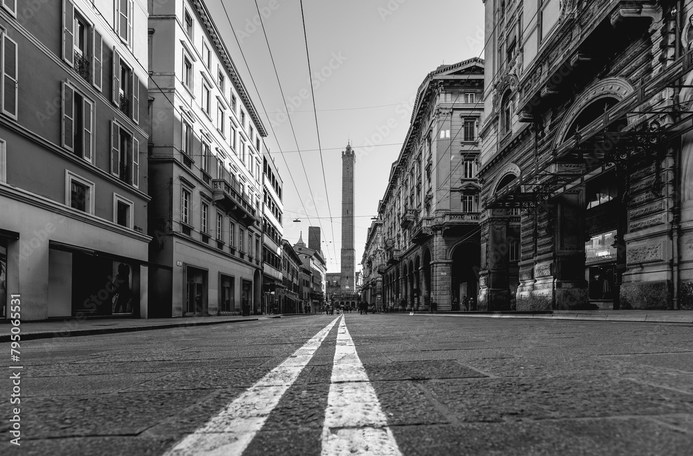 Monochrome image of a Bologna street taken from a low angle, focusing on the converging road lines that lead toward the towering Asinelli Towers in the background