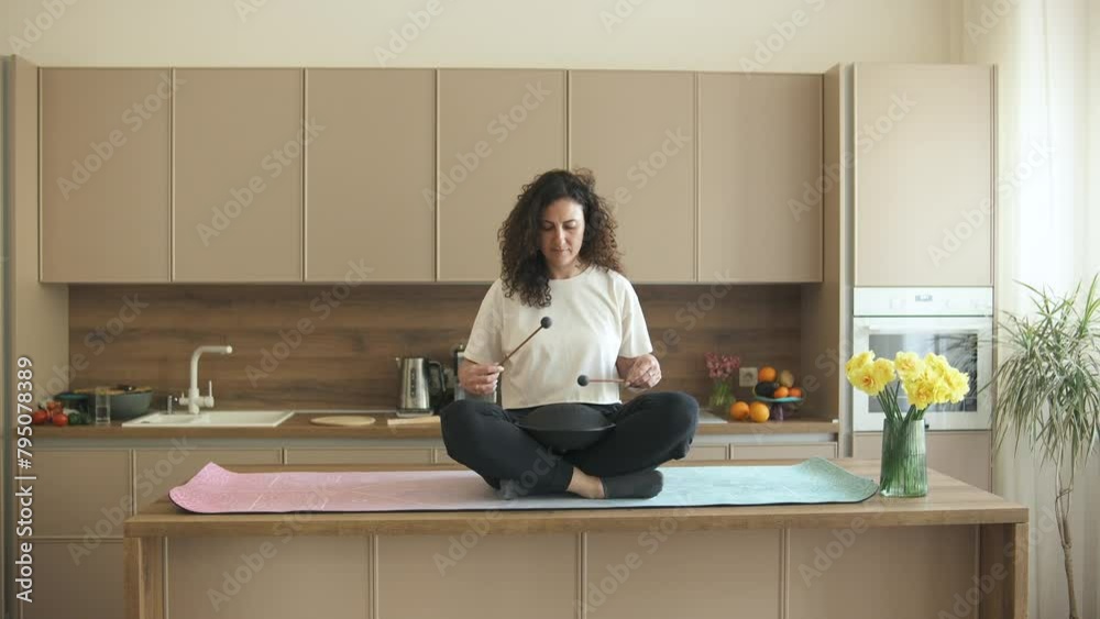 Relaxed curly brunette sitting in lotus pose on the kitchen table and playing tank drum