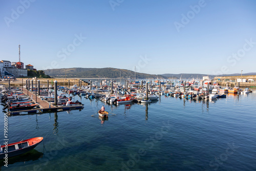 A serene view of a busy harbor in Fisterra Spain filled with various boats, flanked by a town, under the vast expanse of a clear blue sky, depicting tranquility and daily life