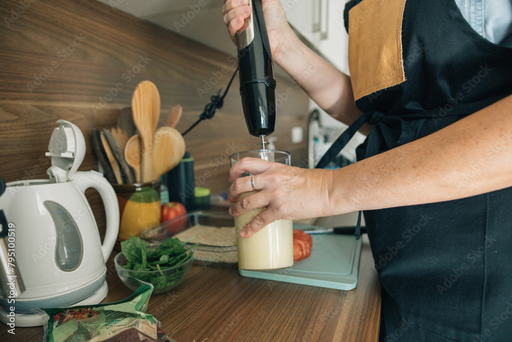A woman is making a smoothie in a kitchen, she is wearing an apron and holding a blender, the kitchen is well-equipped with various utensils and appliances